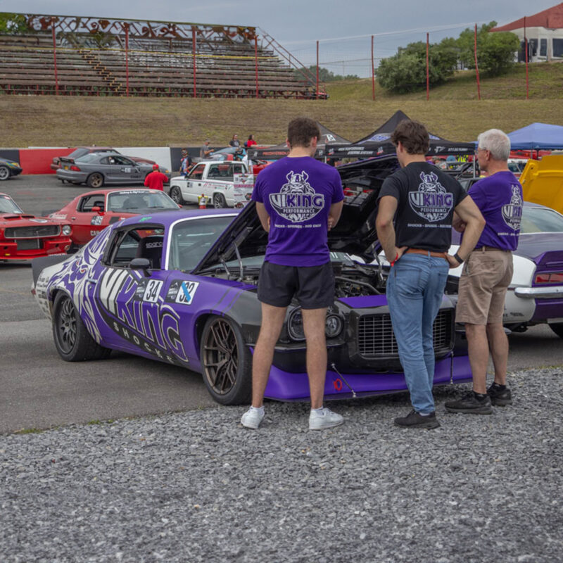 Three men in purple shirts stand in front of a purple race car with its hood open at a racetrack. The car has "Viking" written on its side. Other cars and spectators are visible in the background.