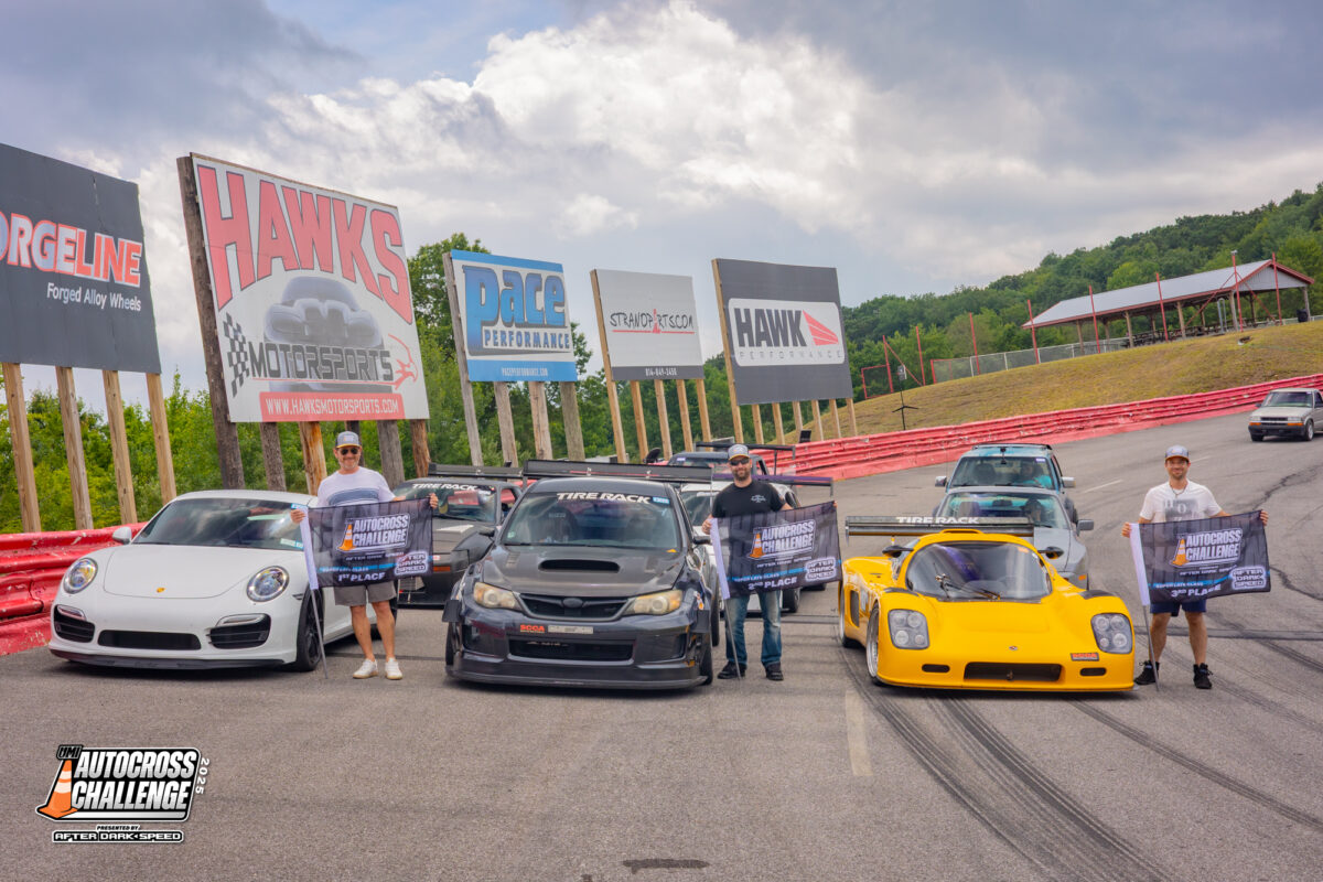 Four people stand holding banners in front of a white Porsche, black Subaru, yellow sports car, and blue car on a racetrack, with sponsor billboards in the background.