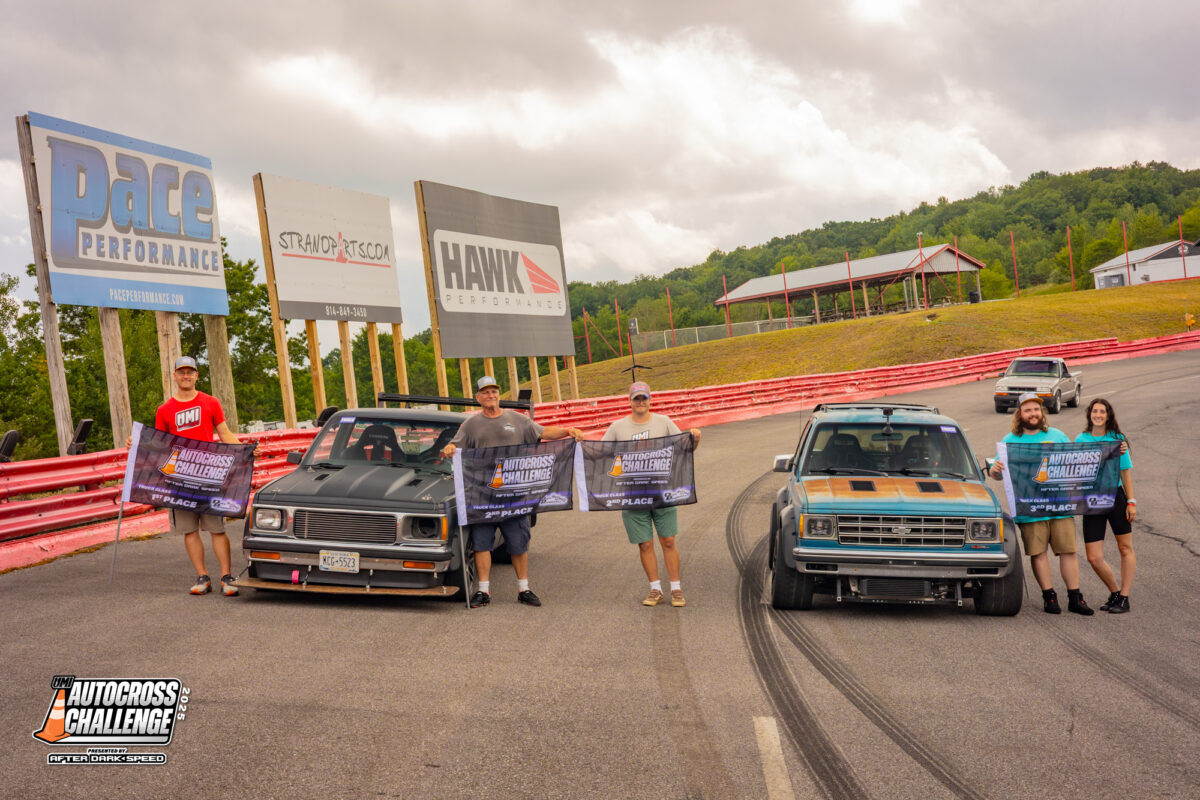 Five people stand next to two vintage trucks on a racetrack, holding banners. Large advertisements are visible in the background on a slightly overcast day.