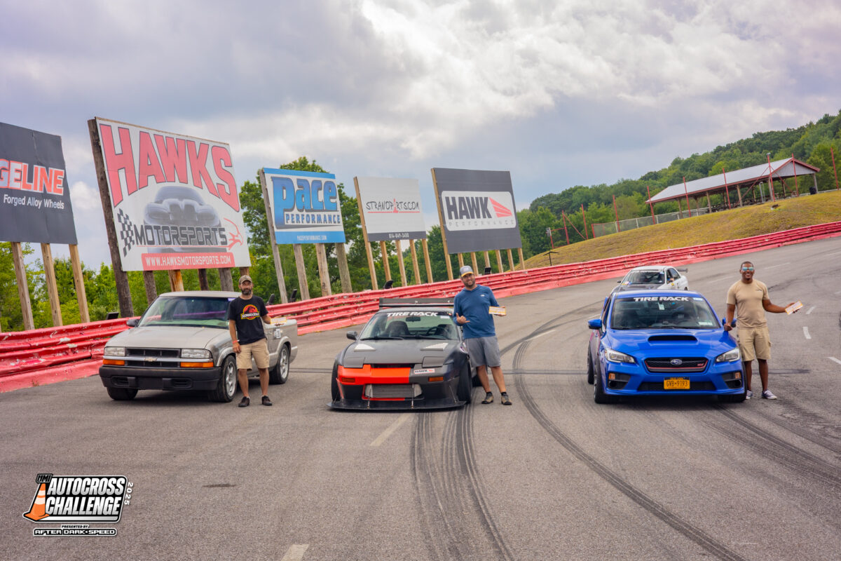 Three men stand beside their modified vehicles on an autocross track, with advertising billboards and green hills visible in the background.