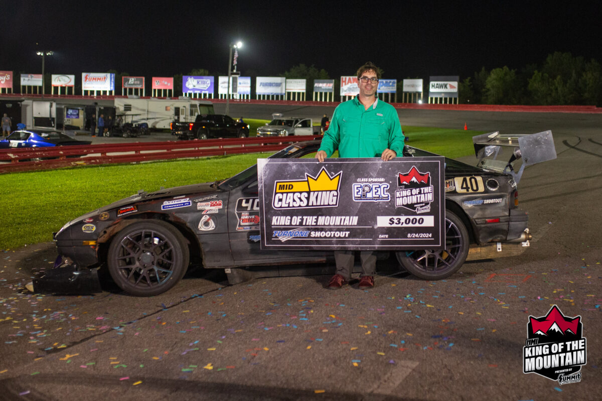 A man in a green shirt holds a large check for $3,000 in front of a race car at a racetrack. The check reads "Mid Class King" and "King of the Mountain." The event banner "King of the Mountain" is visible.