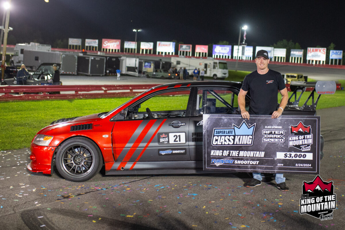 A man in a black shirt and cap stands beside a red racing car holding a large check for $3,000. The car has the number 21 on its door and "King of the Mountain" banners are visible.