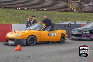 A man is standing next to an orange sports car at a race track.